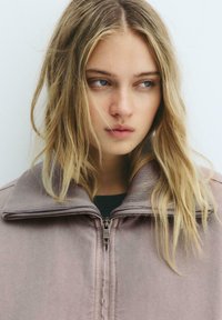 Lys lilla zip-up jakke med en bred ribbet krave. Stoffet ser blødt og struktureret ud, med en subtil glans. Standard lynlås lukning synlig.