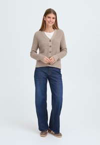 Beige knitted cardigan with a v-neck, five wooden buttons, ribbed texture, worn over a white shirt; paired with wide-leg dark jeans.