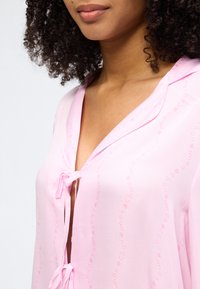 Γυναίκα με σγουρά μαλλιά που φοράει ένα ανοιχτό ροζ μπλούζα δεμένη με δύο φιόγκους στο μπροστινό μέρος, που δείχνει την κλείδ α και το πάνω μέρος του στήθους.