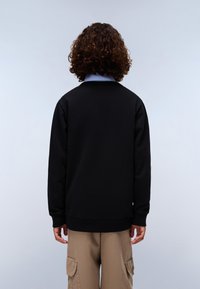 Felpa nera in pile con scollatura rotonda, polsini e orlo a costine. Indossata sopra una camicia a colletto azzurro chiaro. Pantaloni cargo beige visibili.