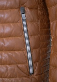 Braune Lederjacke mit gestepptem Muster, ausgestattet mit einem silbernen Reißverschluss auf der linken Seite, hervorgehobenen Nähten und glatter Oberfläche.