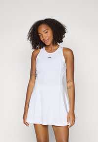 Moteris su garbanotais plaukais, vilkinti be rankovių baltą suknelę su mažu juodu logotipu ant krūtinės, stovi prieš paprastą šviesią fono.