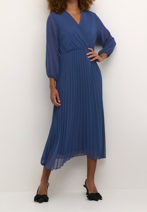 Plisseret midikjole i dybblå stof med indpakningsdesign, lange gennemsigtige ærmer og en flydende silhuet. Båret med sorte hæle med sløjfedetaljer.