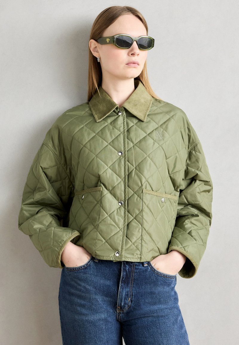 Jachetă matlasată de culoare verde măslinie cu guler din catifea, nasturi tip capcană și buzunare aplicate, purtată cu blugi din denim albastri.