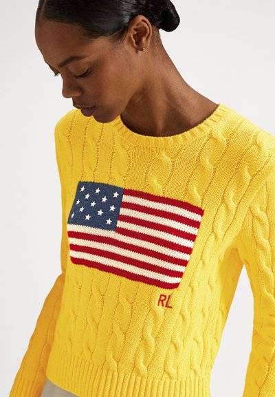 Rumeno pulover z vzorcem pletenega ameriškega zastave na sprednji strani, z rdečimi poudarki ter rebrastimi manšetami in spodnjim robom.