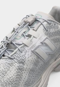 Λευκό αθλητικό παπούτσι με υφασμάτινο επάνω μέρος, ελαστικά κορδόνια χωρίς δέσιμο που συγκρατούνται με πλαστικό κουμπί και μεγάλο λογότυπο "N" στο πλάι.