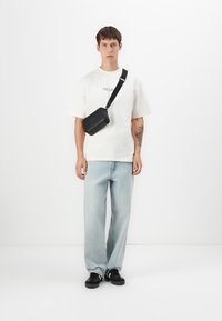 Young man standing wearing a white oversized t-shirt, light blue jeans, black crossbody bag, black sneakers, and a tattoo on his right forearm.