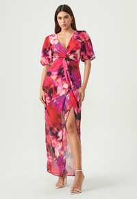 Bloemen maxi-jurk met een diepe V-hals, pofmouwen, een knoopdetail op de taille en een hoge split aan de zijkant; voornamelijk in roze en paarse tinten.