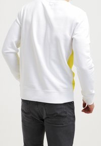 Sudadera blanca con paneles laterales amarillos, cuello redondo y mangas largas. El tejido parece suave; cuenta con un dobladillo recto y puños acanalados.