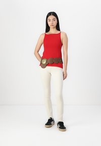 Top en maille rouge à côtes avec une ceinture marron ornée d'accents ronds marron et doré, associé à un jean skinny couleur crème et des baskets noires.