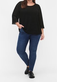 Personne portant un haut noir texturé à manches trois quarts, un jean skinny bleu foncé et des mocassins noirs brillants, debout avec une main dans la poche.