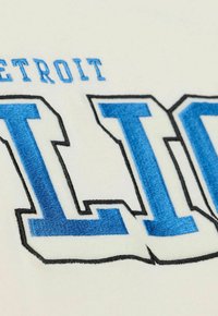 White fabric features a blue embroidered logo reading "DETROIT LIONS," with black outlines highlighting the letters' edges.