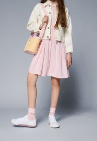 Světle růžové pruhované šaty se zapínáním na knoflíky, v kombinaci s béžovou denimovou bundou a bílými teniskami. Doplňkem je malá pletená kabela ve tvaru kbelíku.