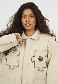 Barna denim dzseki, amelynek zsebei puzzle darabok mintájával díszítettek, barna varrással és egy hímzett folttal, mely egy profil sziluettet ábrázol.