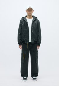 Crna kapuljača jakna s rebrastim manžetama, prebačena preko bijele majice. Tamno sivi cargo hlače imaju više džepova i žuti patentni zatvarač.