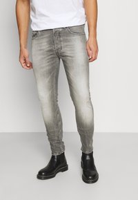 Muž v vybledlých šedých slim-fit džínech a černých kožených Chelsea botách stojící na čistém bílém pozadí.