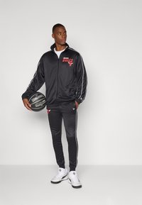 Black athletic track jacket and pants with red Chicago Bulls logo, white stripes, and smooth fabric; holding a Spalding basketball.