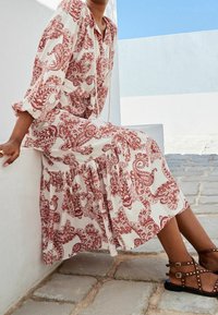 Długa sukienka w beżowym kolorze z czerwonym wzorem paisley, z długimi rękawami i marszczonym dołem. Całość dopełniają czarne sandały na pasku z ćwiekami.