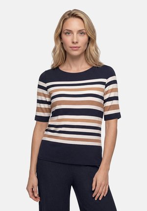 Femme blonde portant un t-shirt à manches courtes avec des rayures horizontales bleu marine, blanc et beige, debout devant un fond clair uni.