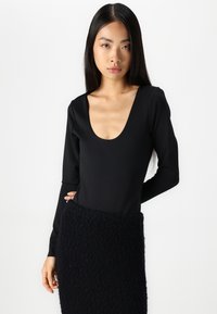 Svart langermet bodysuit med dyp utringning, kombinert med et strukturert mørkt skjørt. Glatt stoff og tettsittende design.