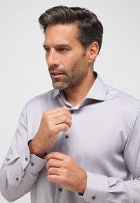 Helle graue Hemd mit subtiler Textur, langen Ärmeln und dunklen Knöpfen, getragen von einem Mann, der seinen Kragen anpasst.