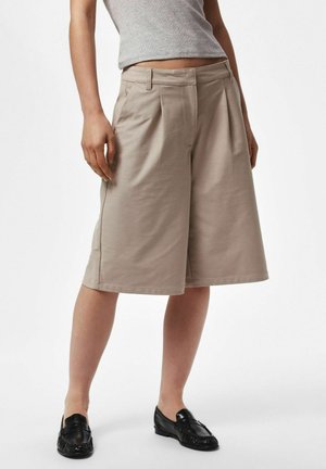 Femme portant un short large beige aux genoux, un top court gris et des mocassins noirs, debout devant un fond blanc.