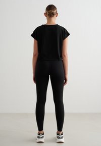 Femme debout de dos, portant un t-shirt court noir, des leggings noirs, des baskets blanches, les cheveux attachés en chignon, sur un fond clair uni.