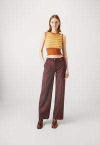 Crochet-knit sleeveless top in orange and yellow, paired with wide-leg maroon trousers. Models brown patterned heeled boots. Minimalist backdrop.