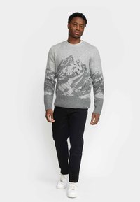 Grauer Strickpullover mit Bergmuster in dunklerem Grau. Rundhalsausschnitt, lange Ärmel, gerippte Bündchen und Saum. Kombiniert mit schwarzen Hosen und weißen Sneakers.