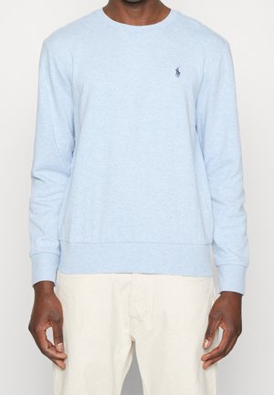 Homme portant une chemise à manches longues bleu clair avec un petit logo de joueur de polo foncé sur la poitrine et un pantalon blanc cassé.