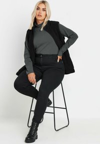 Czarne, wysokogatunkowe dżinsy o gładkiej fakturze; zestawione z ciemnoszarym, długim rękawem topem oraz czarną, puszystą kamizelką. Modelka siedzi na czarnym stołku.