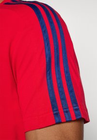T-shirt de sport rouge avec un motif à trois rayures bleues sur les épaules. Texture côtelée avec un ourlet droit et des manches courtes.