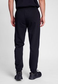 Schwarze Jogginghose mit elastischem Bund und Bündchen, aus weichem Material. Das Modell trägt schwarze Sportschuhe mit strukturierten Details.
