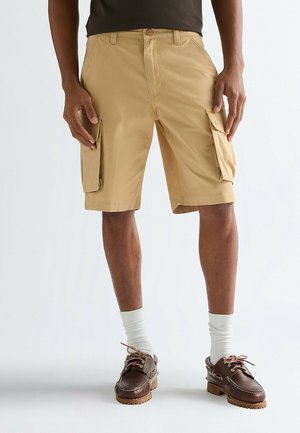Beige Cargo-Shorts mit zwei Seitentaschen, leichtem Stoff und geradem Schnitt. Kombiniert mit weißen Socken und braunen Bootsschuhen.
