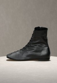 Μαύρο δερμάτινο μποτάκι αστραγάλου με μπροστινά κουμπιά, επίπεδη σόλα και ελαφρώς μυτερή μύτη, τοποθετημένο σε φωτεινή επιφάνεια μπροστά σε γκρι φόντο.