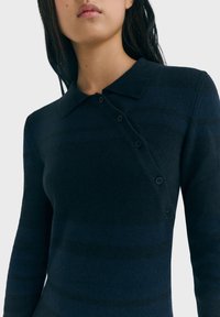 Tummansininen pitkähihaisen neulepolo, jossa on kauluspäällinen, pystysuorat raidat ja kuusi nappia sivussa kiinnitystä varten.