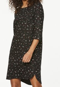 Czarna sukienka o luźnym kroju, z rękawami 3/4, z kolorowym abstrakcyjnym wzorem białych, czerwonych i niebieskich kształtów. Widziane są wyraźne rozcięcia boczne.