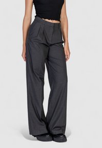 Pantaloni ampi grigi con pieghe, realizzati in tessuto texturizzato. Presentano una vita alta e una vestibilità comoda, abbinati a scarpe nere spesse.