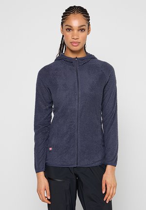 Młoda kobieta z warkoczami, ubrana w granatową bluzę polarową na zamek i czarne spodnie, stoi na jasnym, gładkim tle.