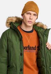 Groene parka met een capuchon met bonttrim, rits- en knoopsluiting, gedragen over een oranje sweatshirt met zwart logo, bedekt met een bruine gebreide beanie.