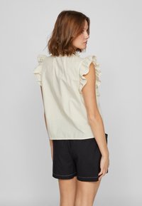 Kremfarget topp med volanger på skuldrene, laget av lett stoff. Brukt med svarte shorts med et enkelt design og kontrasterende søm.