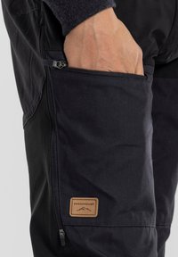 Black fabric pants with a side zip pocket. Features a leather patch with “SWEDEMOUNT” logo. Smooth texture, tapered design.