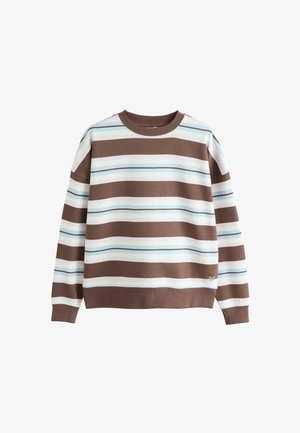 Kvinde iført løse brune bukser, beige sneakers og en hvid og brun langærmet trøje med vandrette striber, stående foran en beige væg.