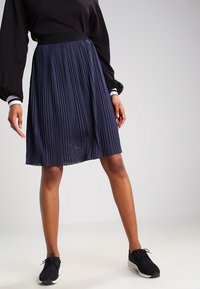 Marinblå plisserad kjol med en slät elastisk midja, som har ett texturerat mönster och en knälång skärning, kombinerad med svarta träningsskor.
