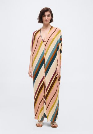 Γυναίκα που φοράει ένα μακρύ, φαρδύ φόρεμα με κουμπιά και πολύχρωμες διαγώνιες ρίγες σε ροζ, κίτρινο, τυρκουάζ και καφέ, στέκεται με ουδέτερη έκφραση.