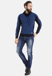 Blau und schwarz gestrickter Pullover mit einem Cowl-Neck, strukturiertem Muster und Kordeln. Tragen mit ausgewaschenen blauen Jeans und schwarzen Stiefeln.