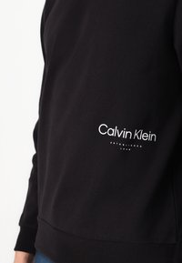 Camisa negra de manga larga con el logo blanco "Calvin Klein Established 1968" en el lado izquierdo del pecho, llevada por una persona con jeans azules visibles por debajo.