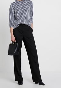 Svarta vida byxor i kombination med en rutmönstrad topp i svart och vitt med trekvartsärmar; bär en svart väska med gulddetaljer.