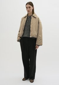 Beige kort bomberjacka med dragkedja, bärs över en grå tröja och svarta högmidjade byxor. Strukturerade svarta skor fulländar looken.