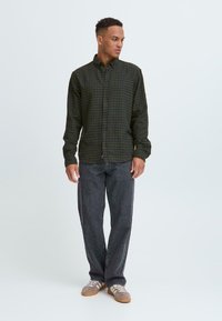 Camisa de botones en cuadros oscuro verde y azul marino, con cuello y mangas largas, combinada con pantalones de mezclilla gris oscuro y zapatillas claras.